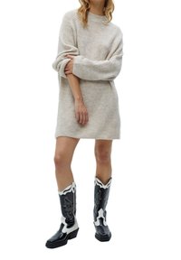 Robe pull beige en tricot avec des manches longues et une coupe ample, associée à des bottes de cowboy noires et blanches avec des embellissements et des détails en patchwork.