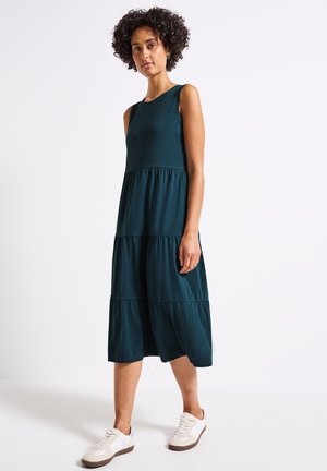 Vestido sin mangas de color verde azulado con diseño en capas, hecho de una tela suave, que cae por debajo de la rodilla, combinado con zapatillas blancas con suelas marrones.