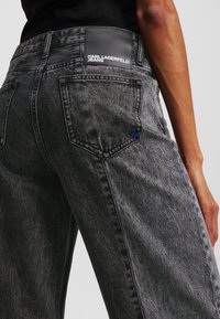 Pilkos džinsinės kelnės su laisvu siluetu, turinčios blizgantį juodą etiketę su inscriptions "KARL LAGERFELD JEANS" ir mažą mėlyną logotipą ant užpakalinio kišenės.