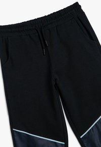 Pantalones de chándal negros hechos de una mezcla de algodón con cintura elástica, que cuentan con bolsillos laterales y un panel brillante en contraste en el dobladillo.