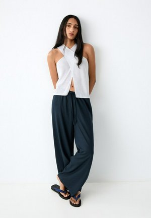 Donna con capelli lunghi e scuri che indossa un top bianco senza maniche, pantaloni neri larghi e infradito blu, in piedi contro un muro bianco.