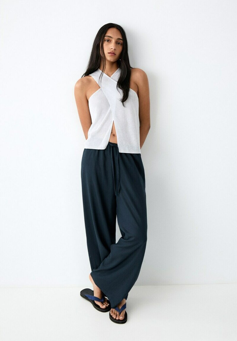 Frau mit langen dunklen Haaren, die ein ärmelloses weißes Oberteil, weite schwarze Hosen und blaue Flip-Flops trägt, steht vor einer weißen Wand.