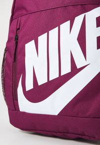 Ljubičasti Nike ruksak sa velikim bijelim logotipom. Izrađen od teksturiranog materijala s bočnom džepnom rajsferšlusom i mrežastim detaljima.