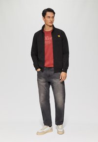 Schwarze Zip-Jacke mit gelbem Logo, rotes Shirt mit weißem Text, graue Jeans, hellfarbige Turnschuhe. Glatter Stoff mit lockerer Passform.