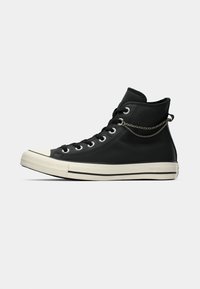 Sneaker alta in pelle nera con suola in gomma bianca, dotata di occhielli metallici, chiusura con lacci e dettaglio in catena sulla caviglia.