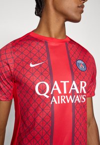 Rød fodboldtrøje med diamantmønster, der har Nike-logo og teksten QATAR AIRWAYS. Designet inkluderer marineblå lodrette striber og et badge.