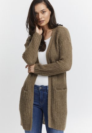 Brun strikket cardigan med et strukturert mønster, åpen front og to lommer foran, kombinert med en hvit topp og blå jeans.