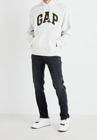 Bílá mikina s kapucí s nápisem "GAP" tmavě zelenou barvou přes hruď, doplněná tmavě šedými džínami a bílými teniskami.