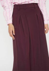 Falda plisada burdeos con un corte ajustado. Textura suave, cinturilla con presillas para cinturón y bolsillos laterales visibles.