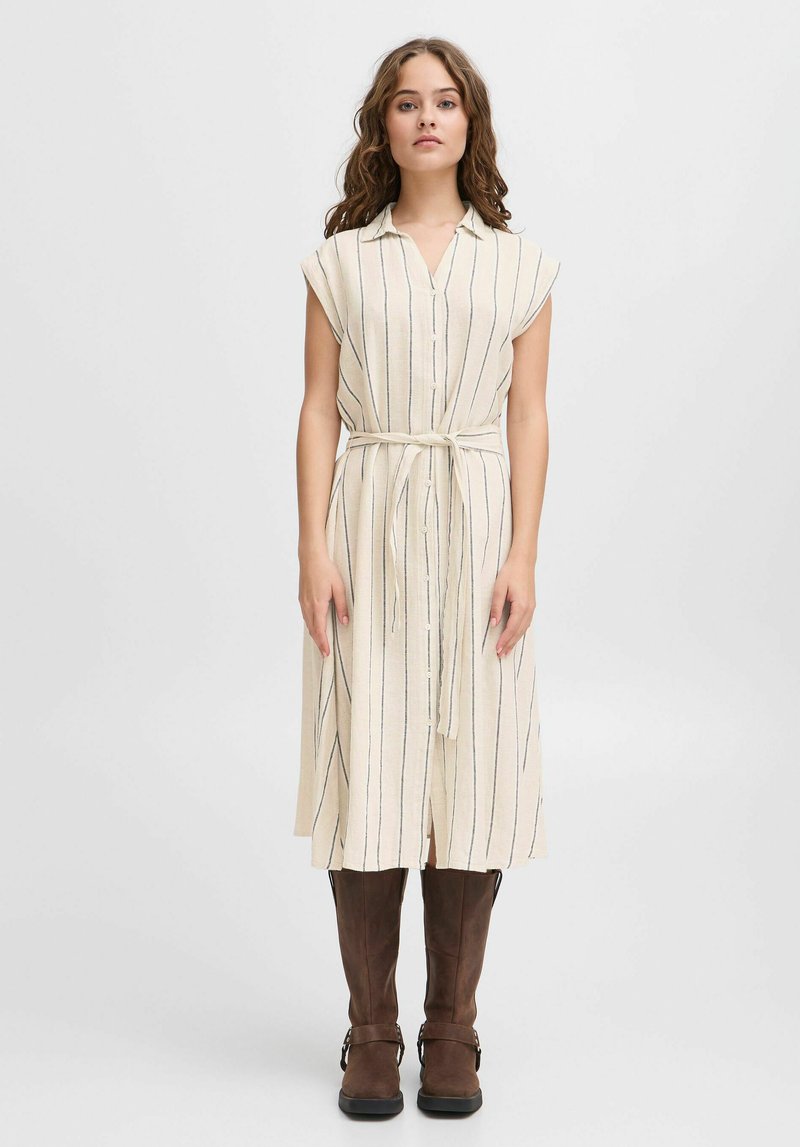Creme-farbenes, knielanges Kleid mit vertikalen marineblauen Streifen, kurzen Ärmeln, durchgehender Knopfleiste und einem gebundenen Taillengürtel, kombiniert mit braunen Stiefeln bis zum Knie.