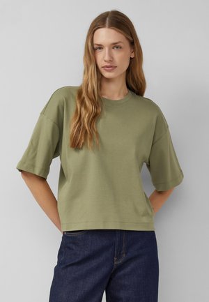 Jeune femme aux longs cheveux blonds portant une chemise oversize manches courtes vert olive et un jean bleu foncé, debout devant un fond gris uni.