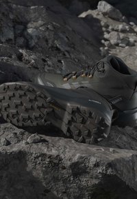 Πολύ σκούρα πράσινα ορειβατικά μποτάκια Adidas Terrex με ανθεκτικές σόλες που ξεκουράζονται σε βραχώδες έδαφος.