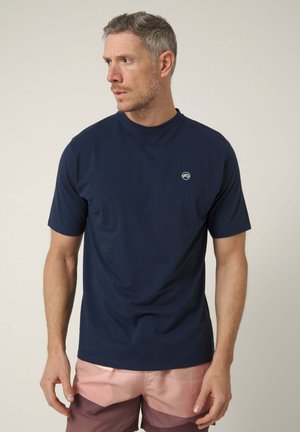 Hombre con el pelo corto, vestido con una camiseta azul marino y pantalones cortos rosas, mirando hacia la izquierda sobre un fondo liso.