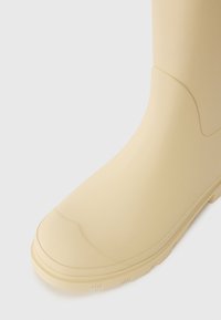 Beige rubber boot with a rounded toe, smooth surface, and textured sole. Features a seamless upper and slight curve near the ankle.