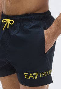 Tengerészkék úszónadrág sárga húzózsinórral és márkajelzéssel. Oldalzsebekkel és könnyű, sima anyagból készült.