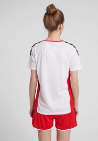 Uniforme deportivo que presenta una parte superior blanca con laterales rojos y rayas negras en los hombros, combinado con pantalones cortos rojos y un escote redondo.
