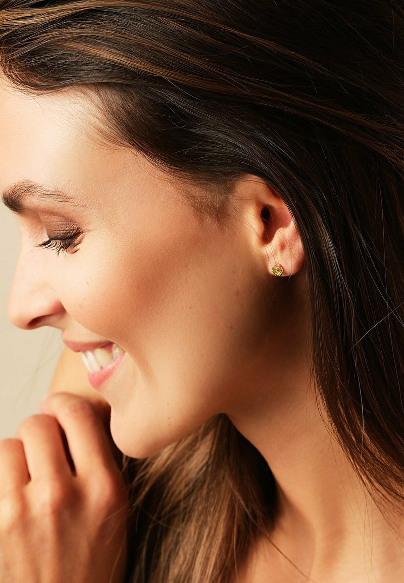 Vrouw met lang bruin haar die zachtjes glimlacht, ogen gesloten, hand bij haar gezicht, met een kleine gouden stud oorbel.