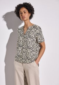 Blusa a maniche corte in crema con motivo astratto nero. Presenta una scollatura a V e una vestibilità ampia, abbinata a pantaloni beige chiaro.