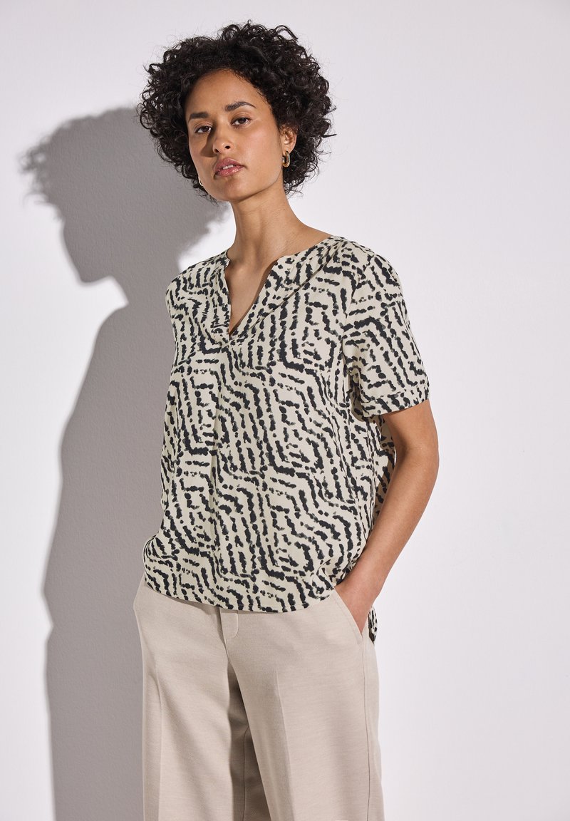 Blusa a maniche corte in crema con motivo astratto nero. Presenta una scollatura a V e una vestibilità ampia, abbinata a pantaloni beige chiaro.
