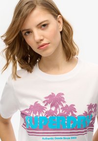 Camiseta de algodón blanca con un gráfico de palmera rosa y texto "SUPERDRY" en turquesa, con cuello redondo y mangas cortas.