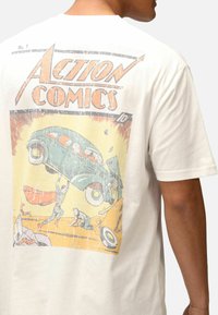 Weißes Baumwoll-T-Shirt mit einem Vintage-Comic-Design auf dem Rücken, das ein grünes Auto zeigt, das angehoben wird, sowie Superheldenfiguren.