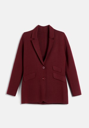 Femme portant un costume bordeaux avec les manches retroussées, un haut blanc, des colliers dorés superposés, une ceinture rouge et des sandales dorées à talons hauts, debout les mains sur les hanches.