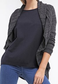 Grauer Strickcardigan mit langen Ärmeln und offenem Front, mit einem strukturierten Design und Fransenakzenten, über einem marineblauen Oberteil und blauen Jeans lagend.