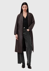 Cappotto lungo in pelle marrone con rever a punta, due tasche frontali, indossato sopra una maglietta nera a coste e jeans neri a vita alta. Stivali neri alla caviglia.