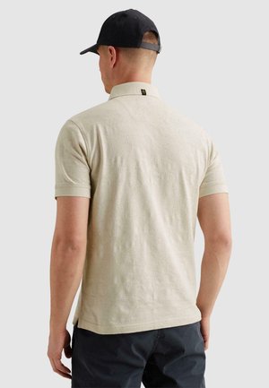 Muž v béžovém texturovaném polokošili s krátkým rukávem a černé čepici, otočený zády k jednoduchému světle šedému pozadí.