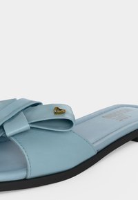 Ανοιχτό μπλε σανδάλι slide με λεπτομέρεια φιόγκου και μικρή χρυσή καρδιά στη μαύρη σόλα, εμφανίζεται από πλάγια γωνία.