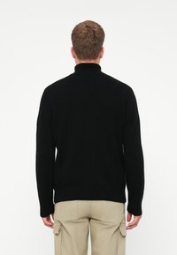 Mies joka seisoo selkä kameraan pukeutuneena mustaan poolopaitaan ja vaaleisiin cargo-housuihin yksinkertaista valkoista taustaa vasten.