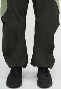Donkergroene broek met een lichtgroene inzet, met een losse pasvorm en zichtbare stikseldetails. Zwarte gestructureerde schoenen met veters onderaan.