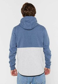 Uomo visto di spalle che indossa un felpa con cappuccio blù e grigio chiaro a blocchi di colore e jeans azzurri su uno sfondo semplice.