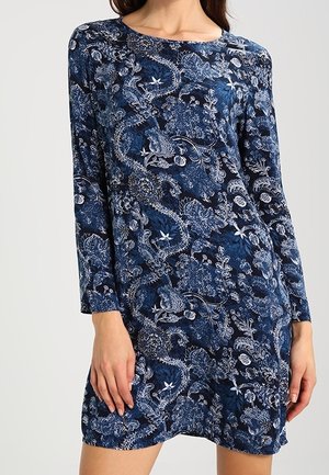 Frau trägt ein langärmeliges, knielanges marineblaues Kleid mit einem kunstvollen weißen Blumen- und Paisleymuster.