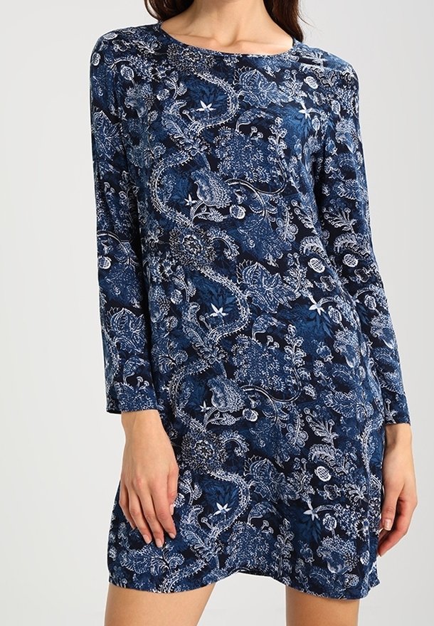Femme portant une robe bleu marine à manches longues, longueur genoux, avec un motif blanc floral et cachemire complexe.
