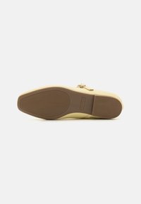 Zapato de lona color beige sin cordones, con una puntera baja y redondeada, parte superior lisa y suela de goma texturizada con surcos horizontales y etiqueta de la marca.