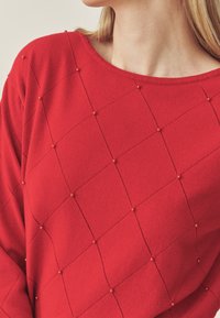 Piros kötött pulóver gyémántmintával és kis gyöngyökkel díszítve. Széles nyakkivágással és texturált anyaggal rendelkezik.