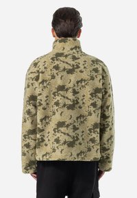Fleecejakke med høj krave, der har et camouflage mønster i olivengrøn og beige. Blød tekstur, brede ærmer og afslappet pasform.
