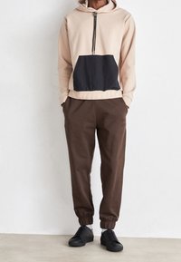 Sudadera beige con un bolsillo frontal negro, cuello con cremallera, combinada con pantalones marrones de cintura elástica y zapatillas negras sobre un fondo neutro.