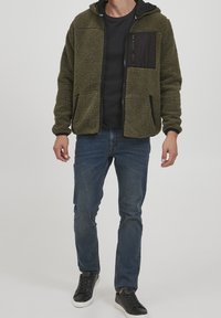 Groene fleece jack met een ritssluiting aan de voorkant, zwarte accenten, twee voorzakken en een capuchon, gecombineerd met donkere jeans en zwarte sneakers.