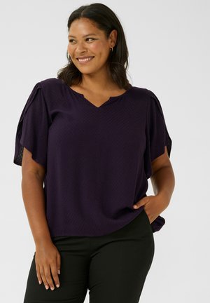 Bluză violet cu mânecă scurtă, decolteu în V și model texturat. Material moale și ușor care se așază delicat pe corp. Se văd pantaloni negri.