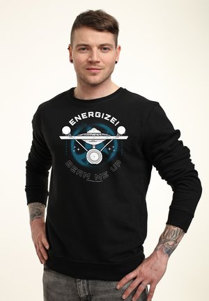 Młody mężczyzna z krótkimi włosami nosi czarną bluzę z grafiką statku kosmicznego i napisem „Energize! Beam me up,” stoi z rękami na biodrach.