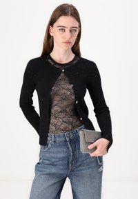 Cardigan nero a coste con dettagli in perle, indossato sopra una blusa in pizzo nero. Abbinato a jeans blu a vita alta e una borsa a tracolla con motivo.