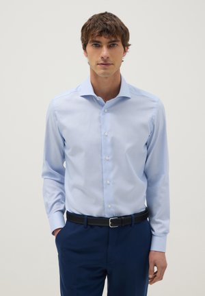 Mann trägt ein hellblaues Hemd mit geknöpftem Kragen und dunkelblaue Hose, steht mit einer Hand in der Tasche vor einem einfarbigen Hintergrund.