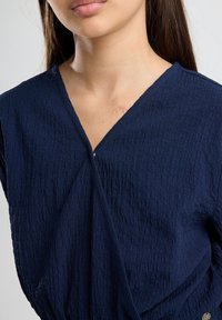 Dark navy blue top with a crinkled texture, V-neckline, and wrapped design, featuring a small metallic accent on the side.