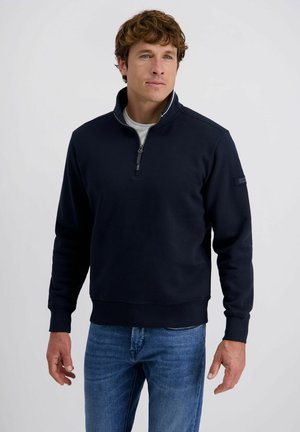 Man wearing a navy quarter-zip pullover over a white shirt and blue jeans, standing against a plain light gray background.