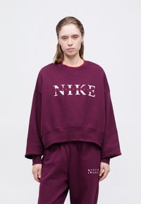 Donna che indossa un completo abbinato di felpa e pantaloni della tuta oversize viola Nike, con loghi bianchi e rossi, in piedi davanti a uno sfondo neutro.