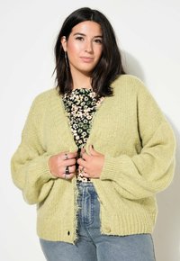 Cárdigan de punto verde claro con un corte holgado, puños acanalados y detalles de dobladillo sin rematar. La modelo lleva una blusa floral debajo.