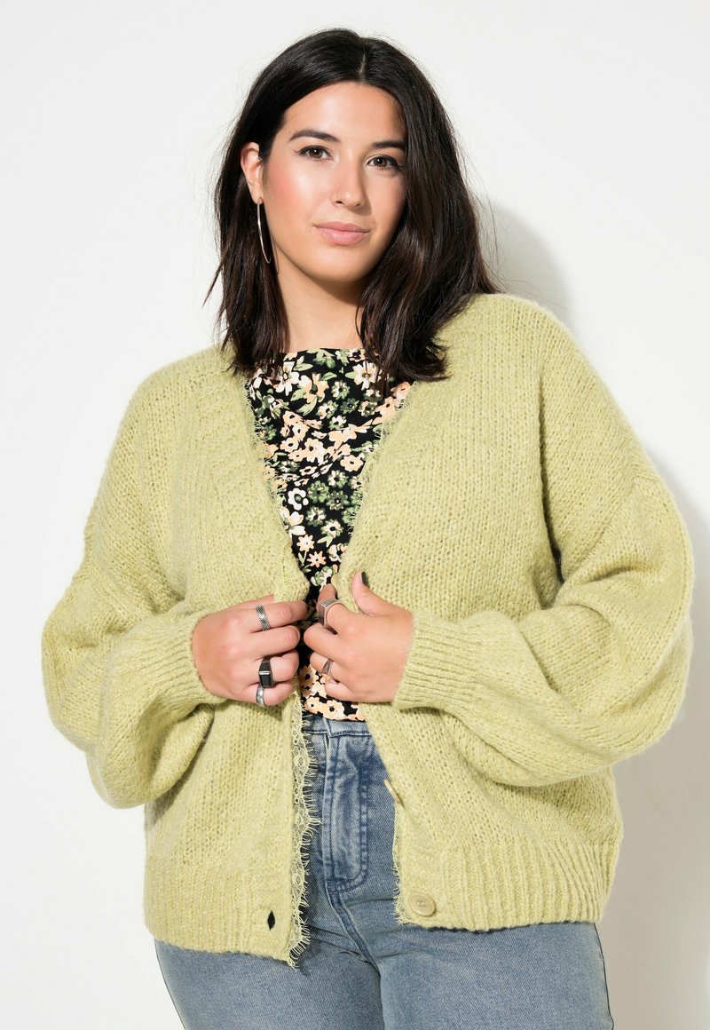 Cárdigan de punto verde claro con un corte holgado, puños acanalados y detalles de dobladillo sin rematar. La modelo lleva una blusa floral debajo.
