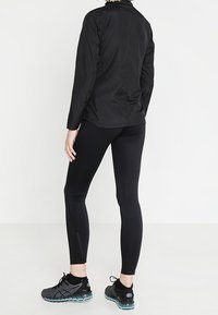 Černá sportovní bunda s vysokým límcem, v kombinaci s přiléhavými černými legínami a černými běžeckými botami s tyrkysovými akcenty, pohled zezadu.